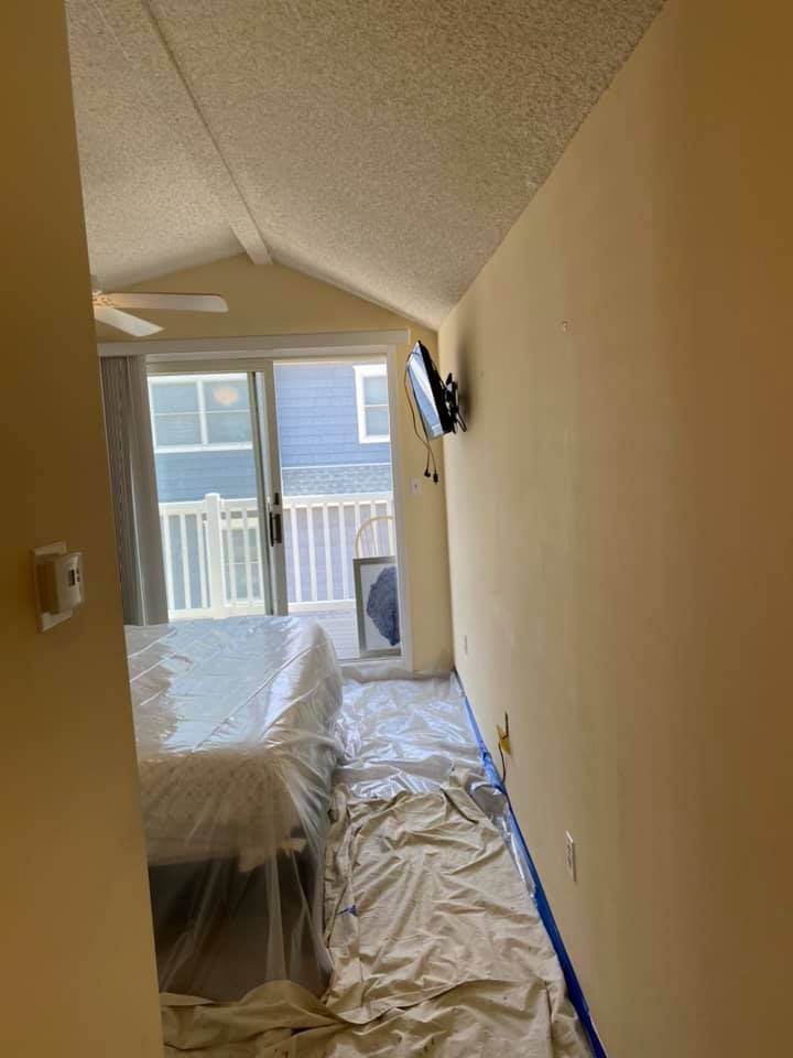 Bedroom with sliding glass door, white bed covered in plastic, beige walls, and a mounted TV.