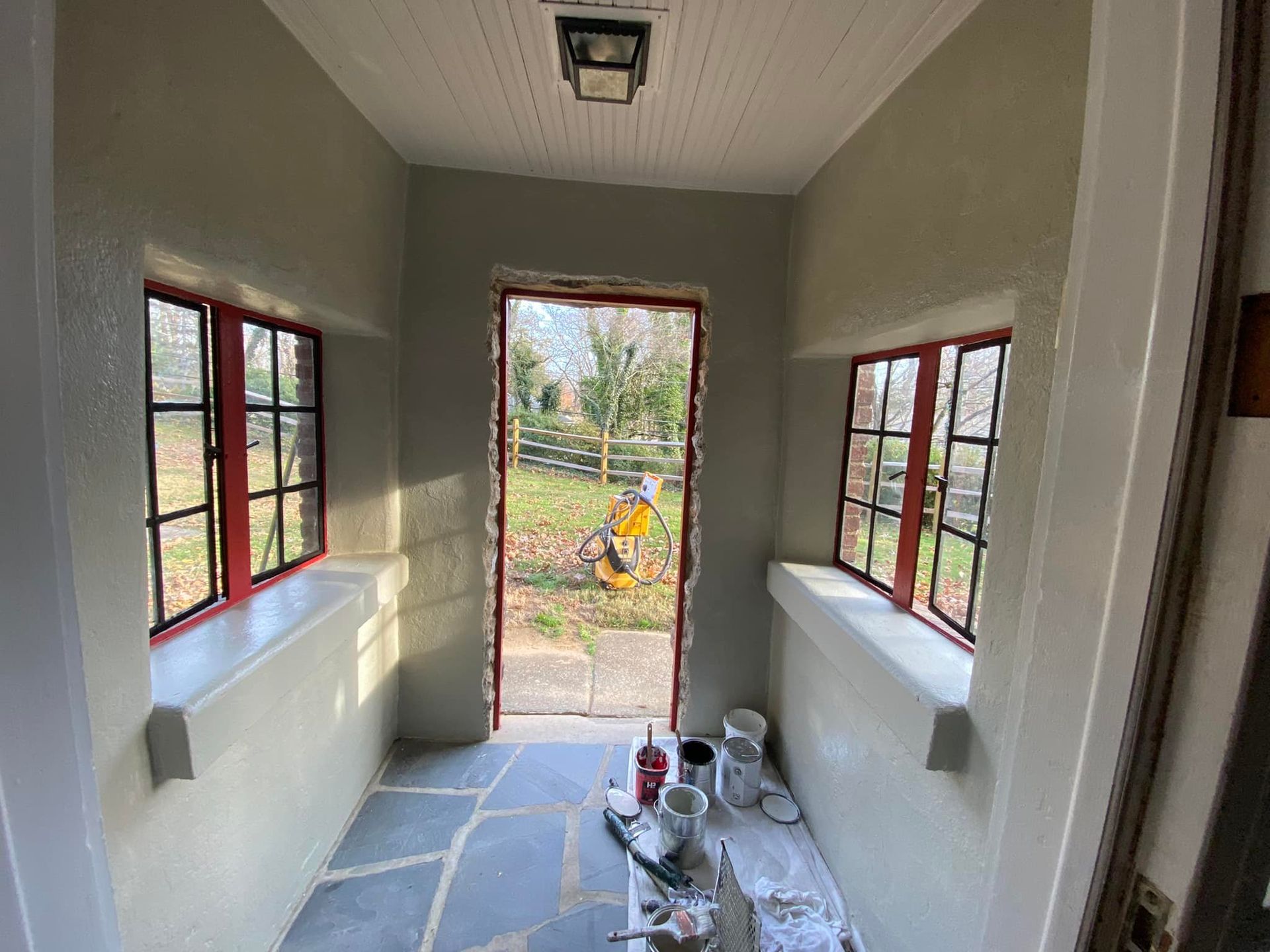 A small room with red-trimmed windows and door, light grey walls, and a stone floor, open to a grassy yard.