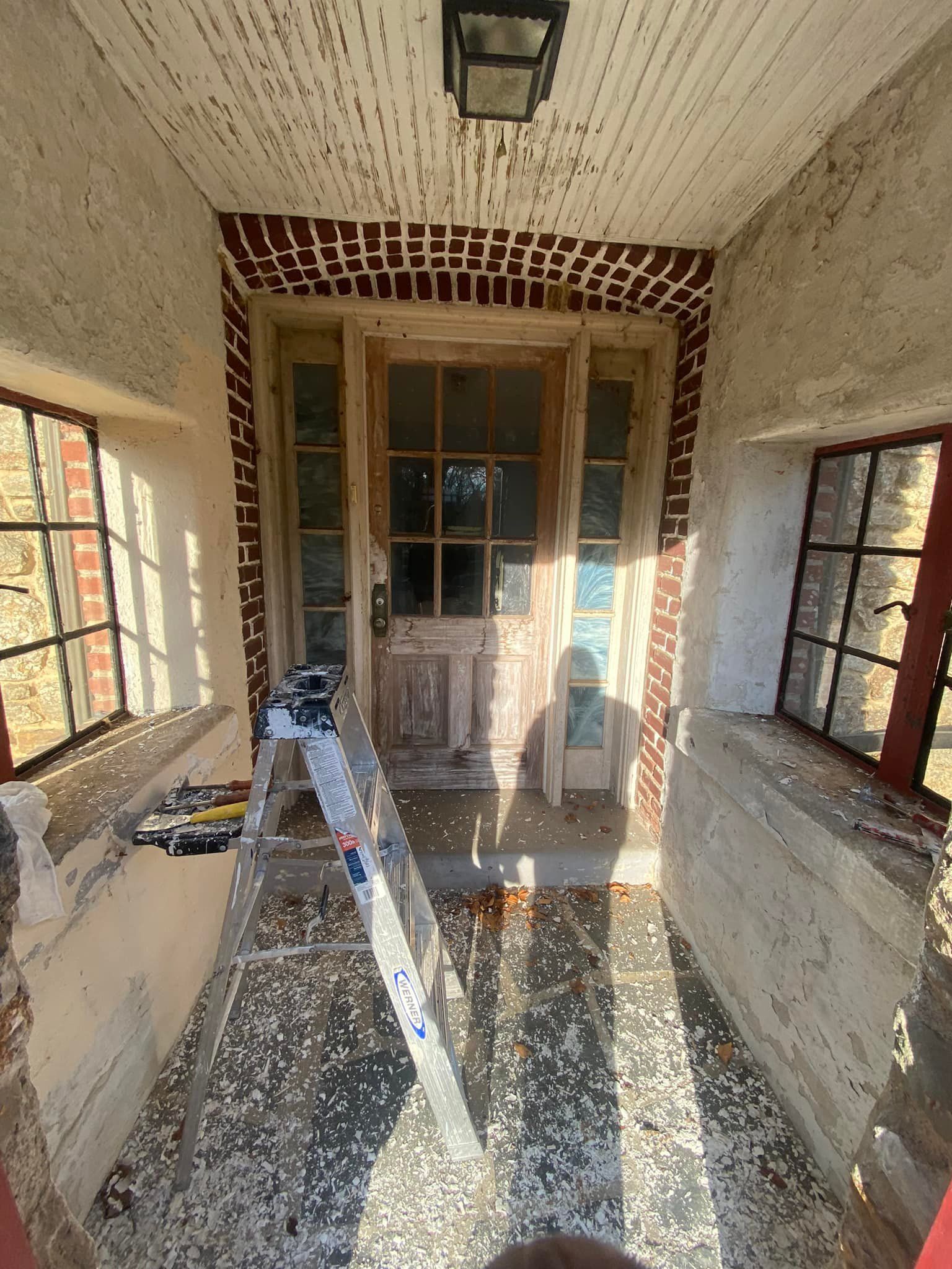 Covered porch with weathered wooden door, windows, and ladder; brickwork and peeling paint.