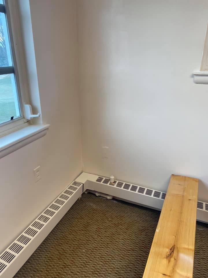 Corner room with window, off-white walls, radiator, and a wooden plank on the floor.