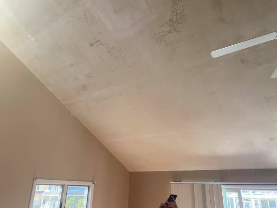 Angled, freshly plastered ceiling in a room with a window, beige walls, and a ceiling fan.