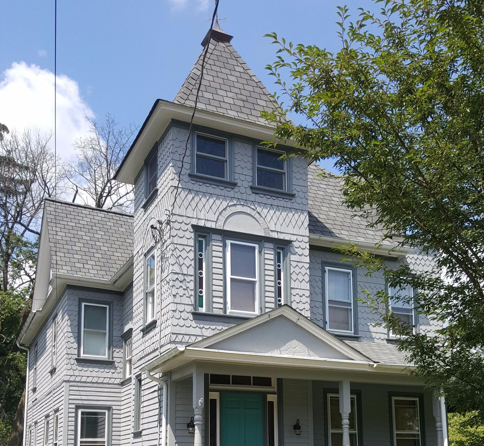 Gray Victorian house with a tower and porch, surrounded by trees under a blue sky.