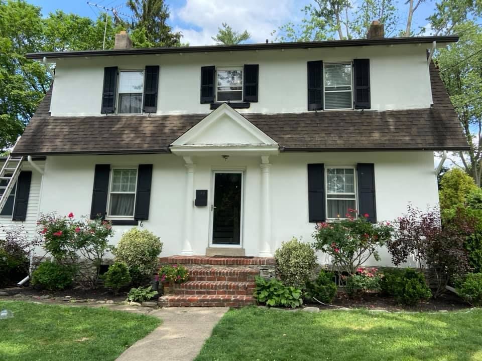 White two-story house with black shutters, brick steps, and a dark roof; roses and bushes in the yard.