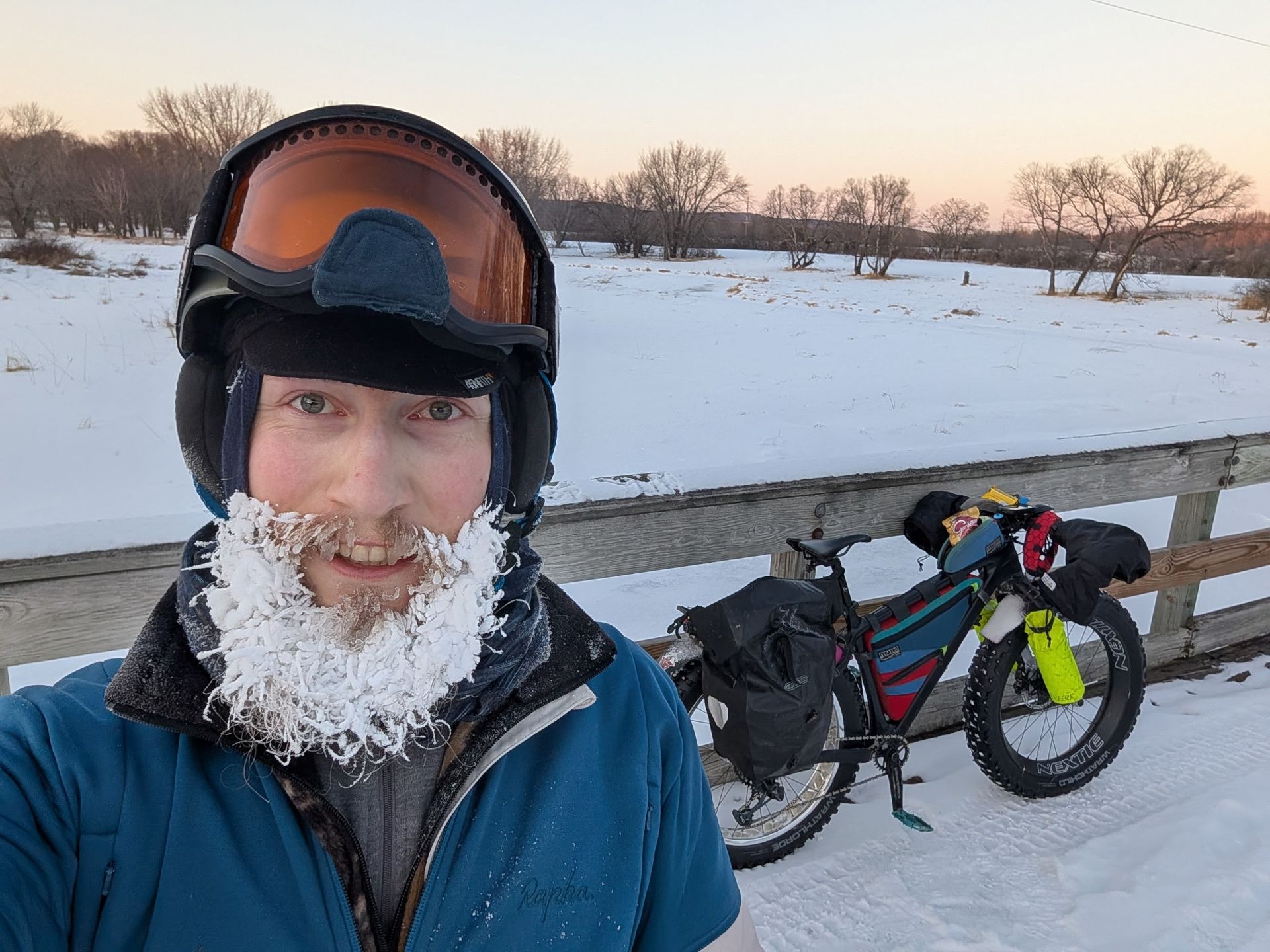 Snowy outdoor selfie of a bundled cyclist beside a loaded bike at sunset