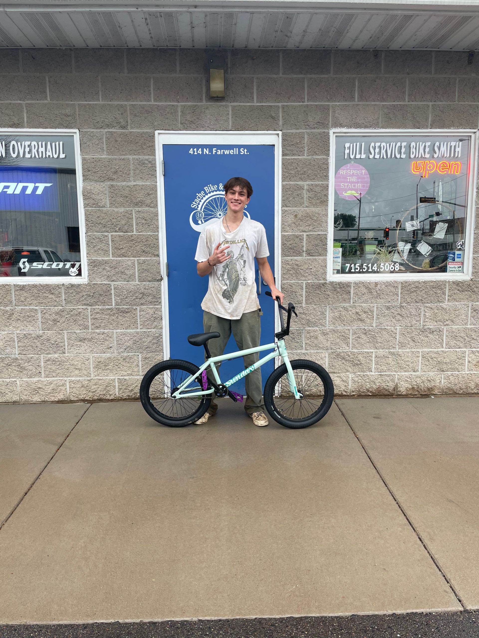 Person standing with a blue-and-black bicycle outside a storefront, holding up a peace sign.