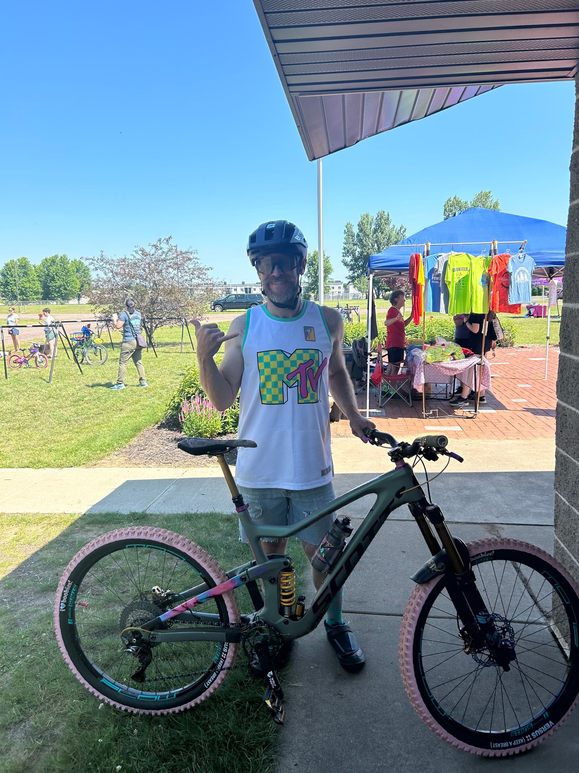 Person standing with a mountain bike at an outdoor event beside colorful tents and people in the background