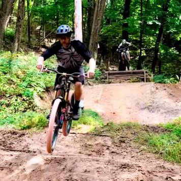 Mountain biker in a blue helmet rides down a muddy forest trail, with another cyclist on a ramp behind.