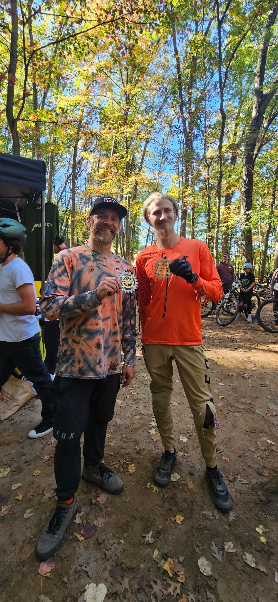 Two people standing on a forest trail, smiling and holding drinks in autumn sunlight.