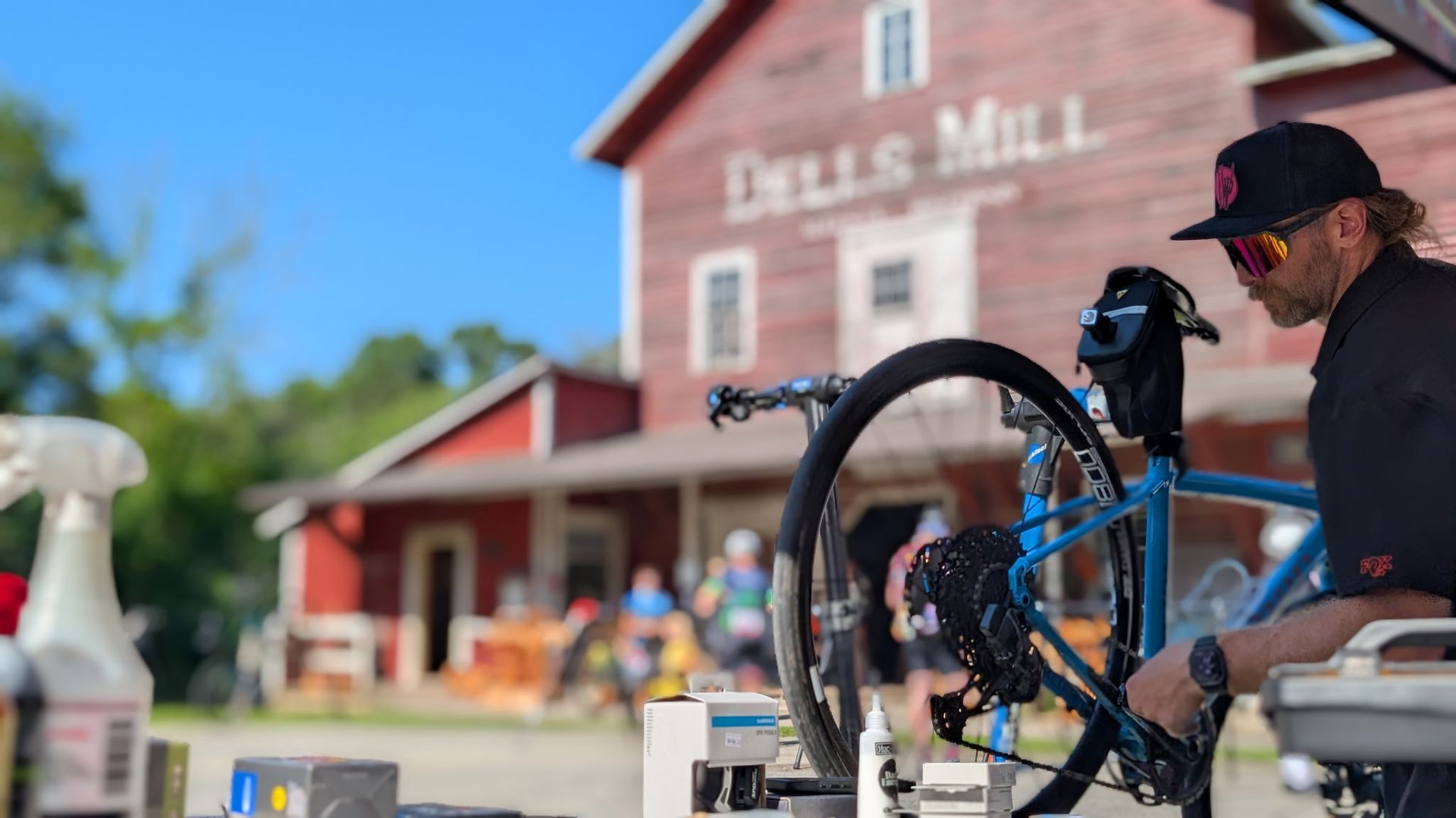 Person fixing a blue bike outside a red mill building on a sunny day