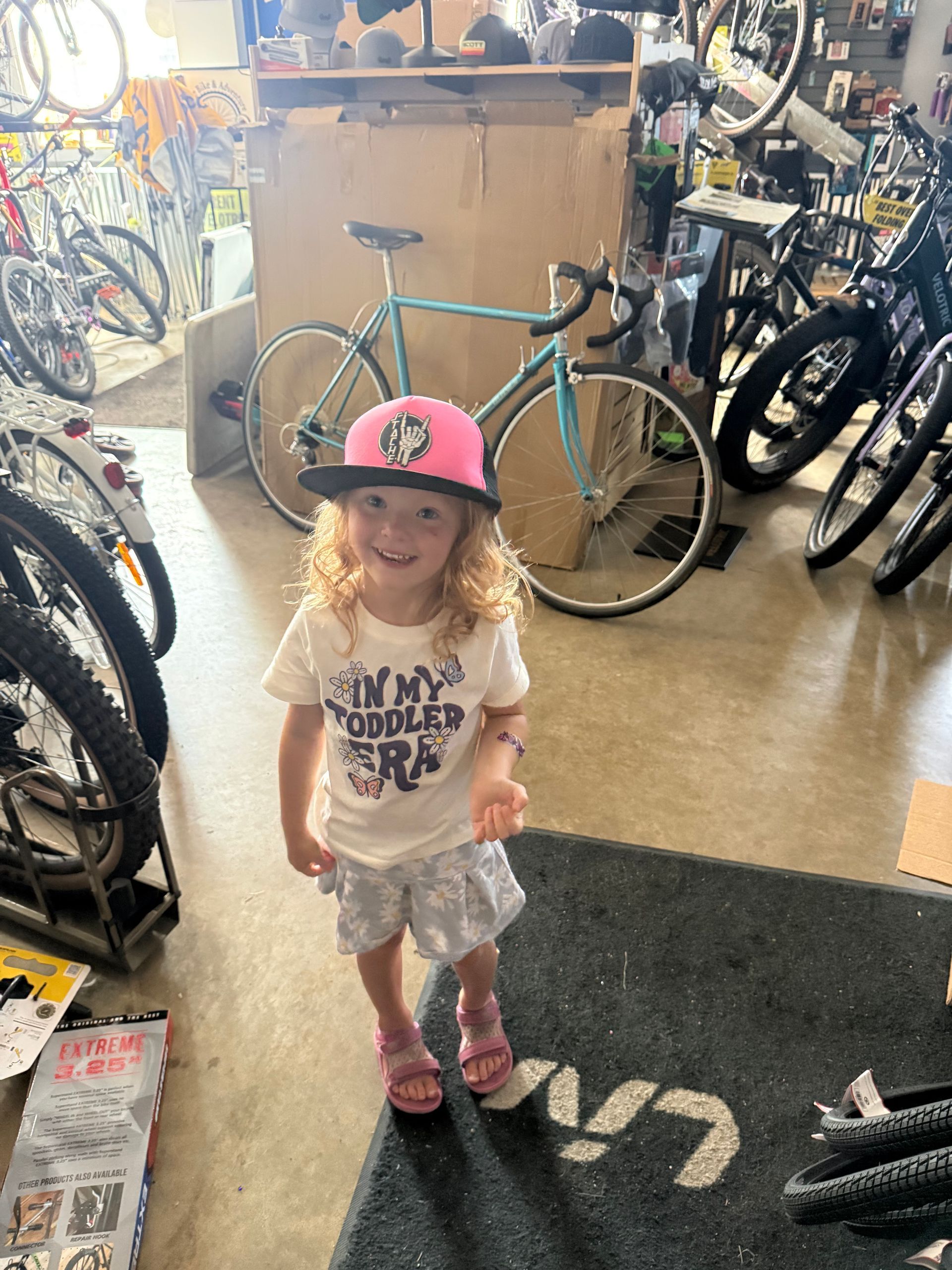 Smiling child in a pink hat standing in a bicycle shop beside a teal bike.