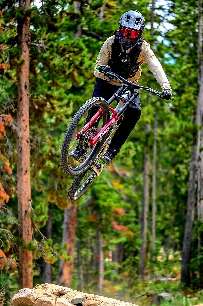 Mountain biker airborne over a forest trail, wearing a helmet and red goggles.