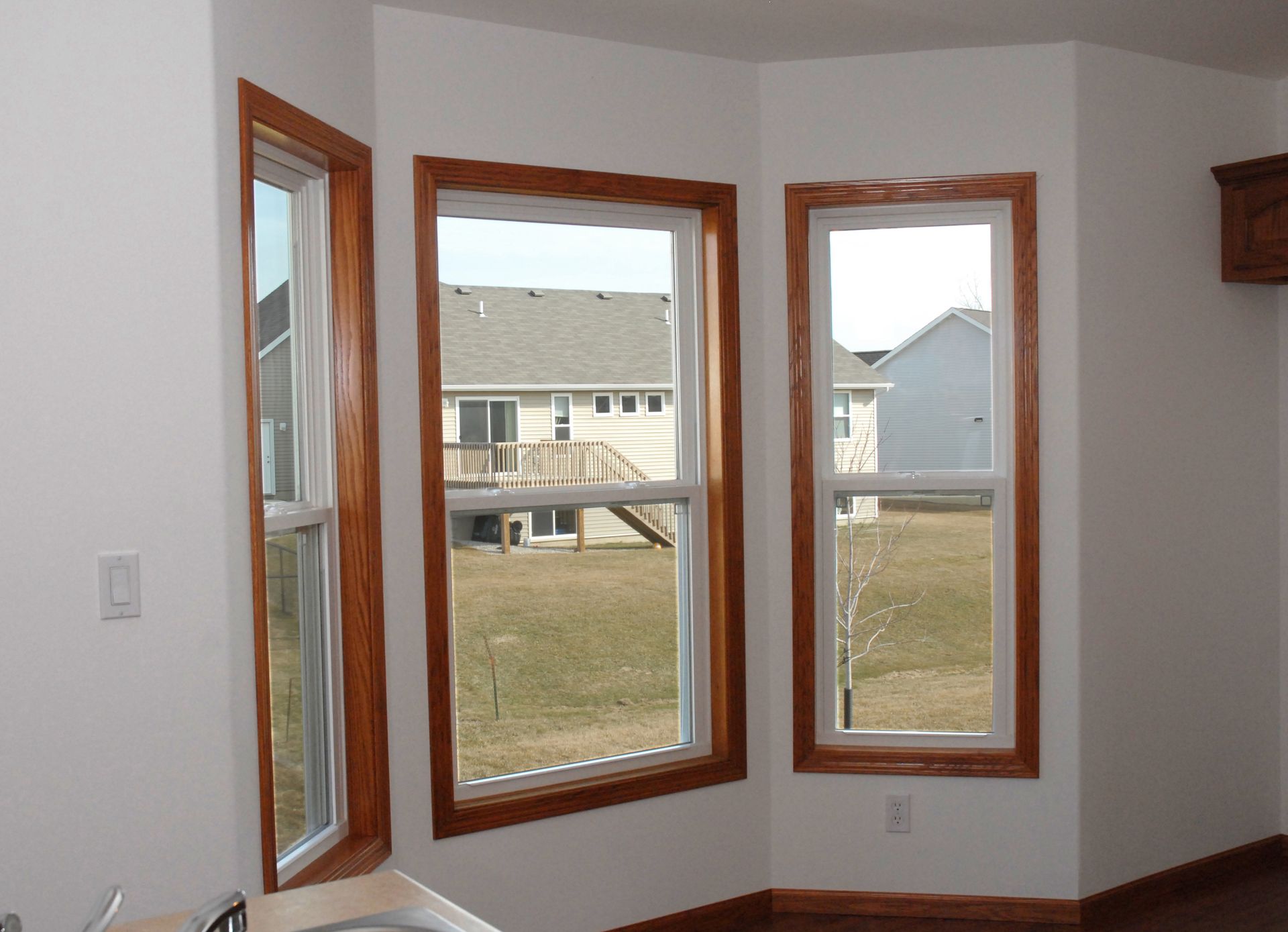 Corner room with three wooden-framed windows overlooking neighboring houses and a lawn.