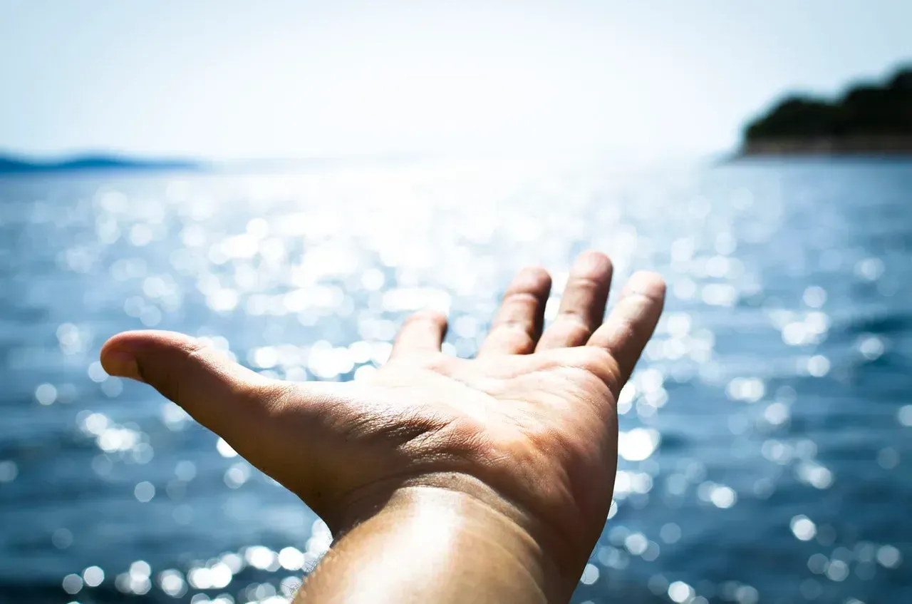 Outstretched hand reaching toward sparkling blue water with a distant shoreline in the background