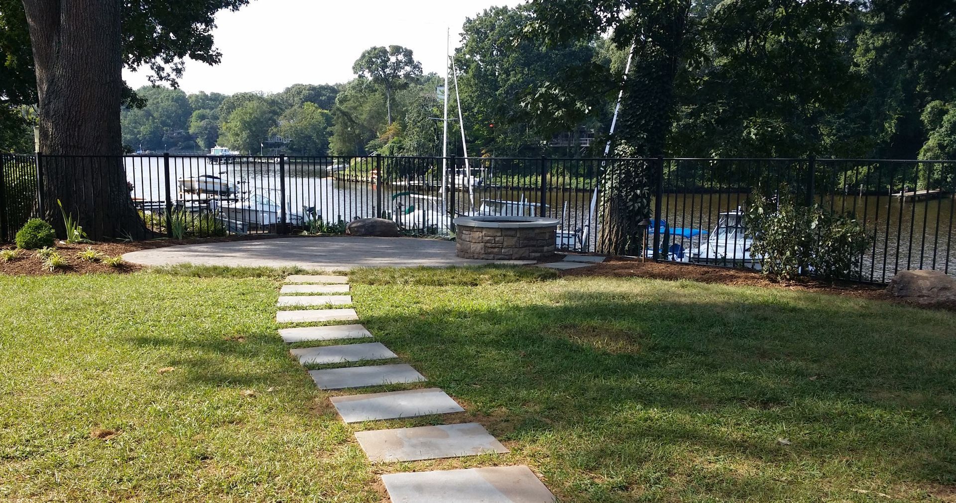 Stone path across a grassy park toward a lake, with trees and a black fence in the background