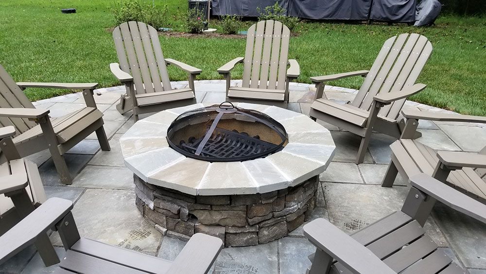 Stone fire pit surrounded by beige Adirondack chairs on a paved patio lawn setting
