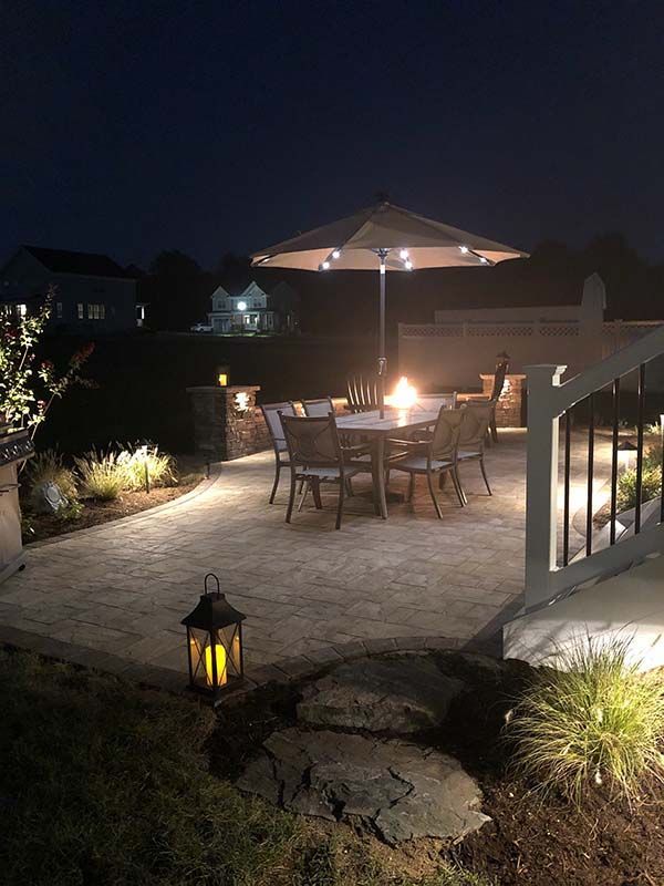 Lit patio at night with table, chairs, umbrella, and lanterns overlooking a backyard garden