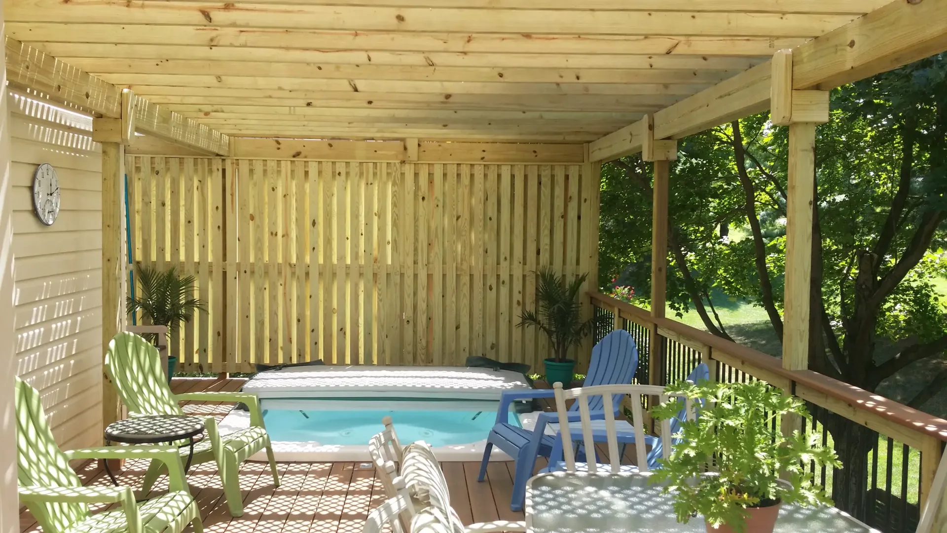 Covered patio with a small hot tub, lounge chairs, potted plants, and wooden railings overlooking greenery