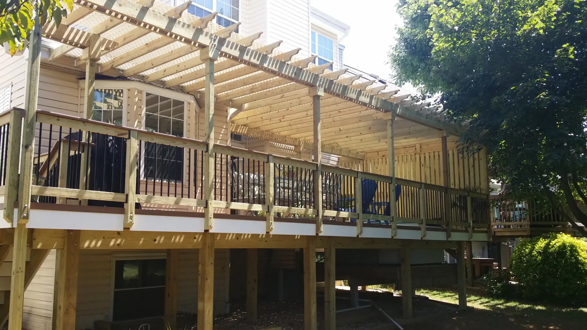 Wooden raised walkway and deck beside a building, with pergola beams and trees in bright sunlight