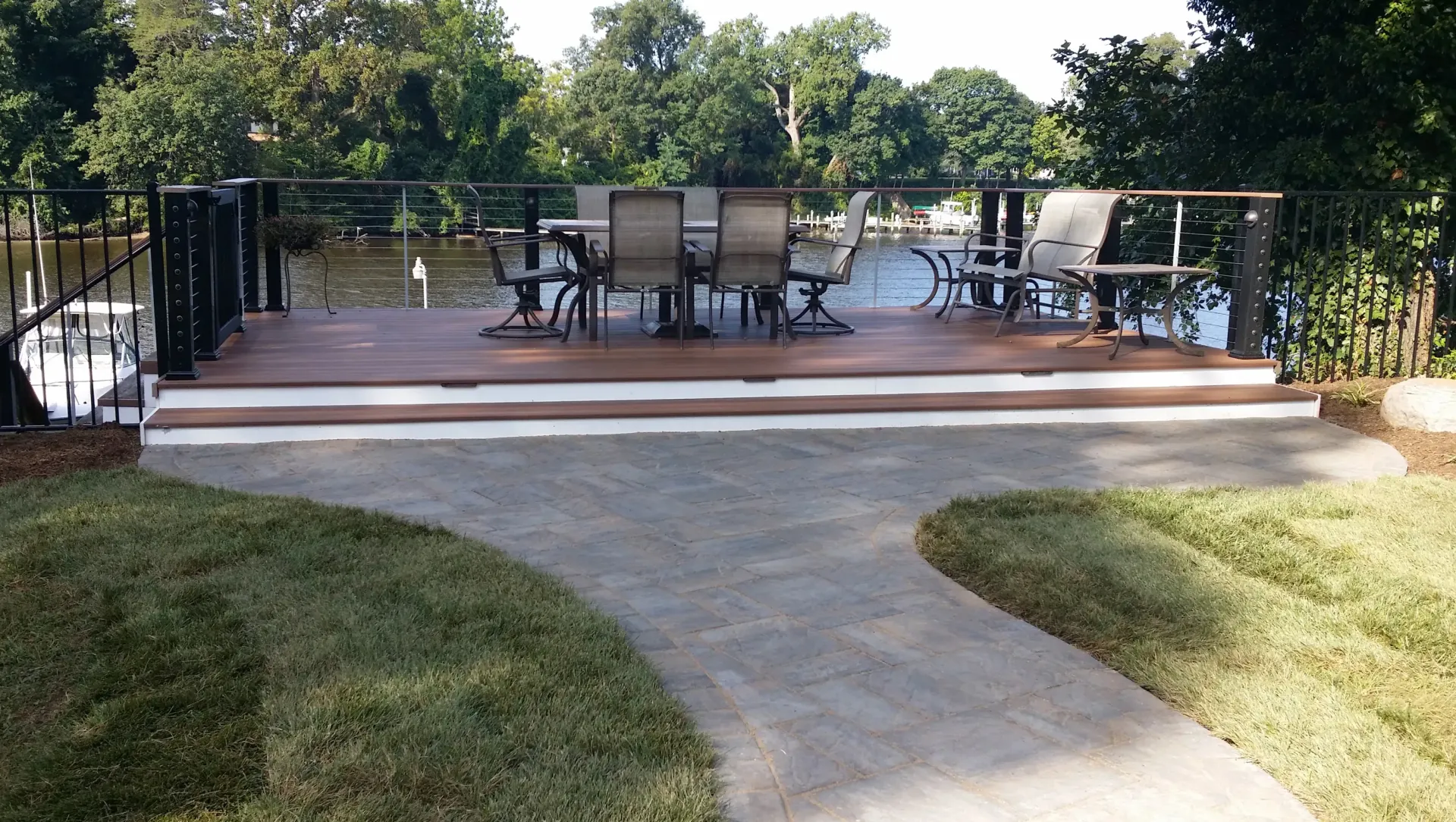 Patio deck with outdoor dining table and chairs beside a curved stone walkway, surrounded by trees.