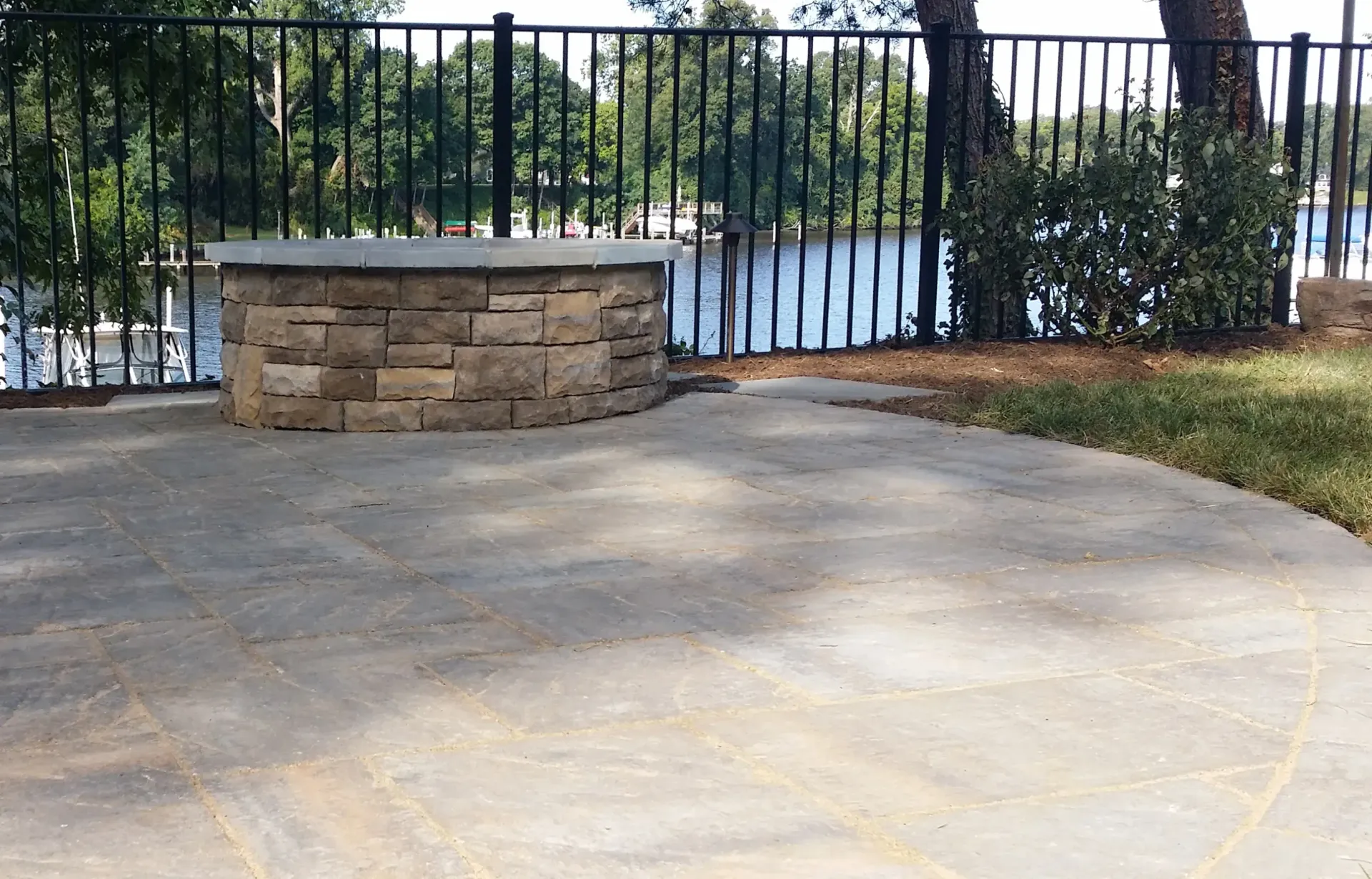 Stone patio beside a black metal fence and lake, with grass and trees in the background