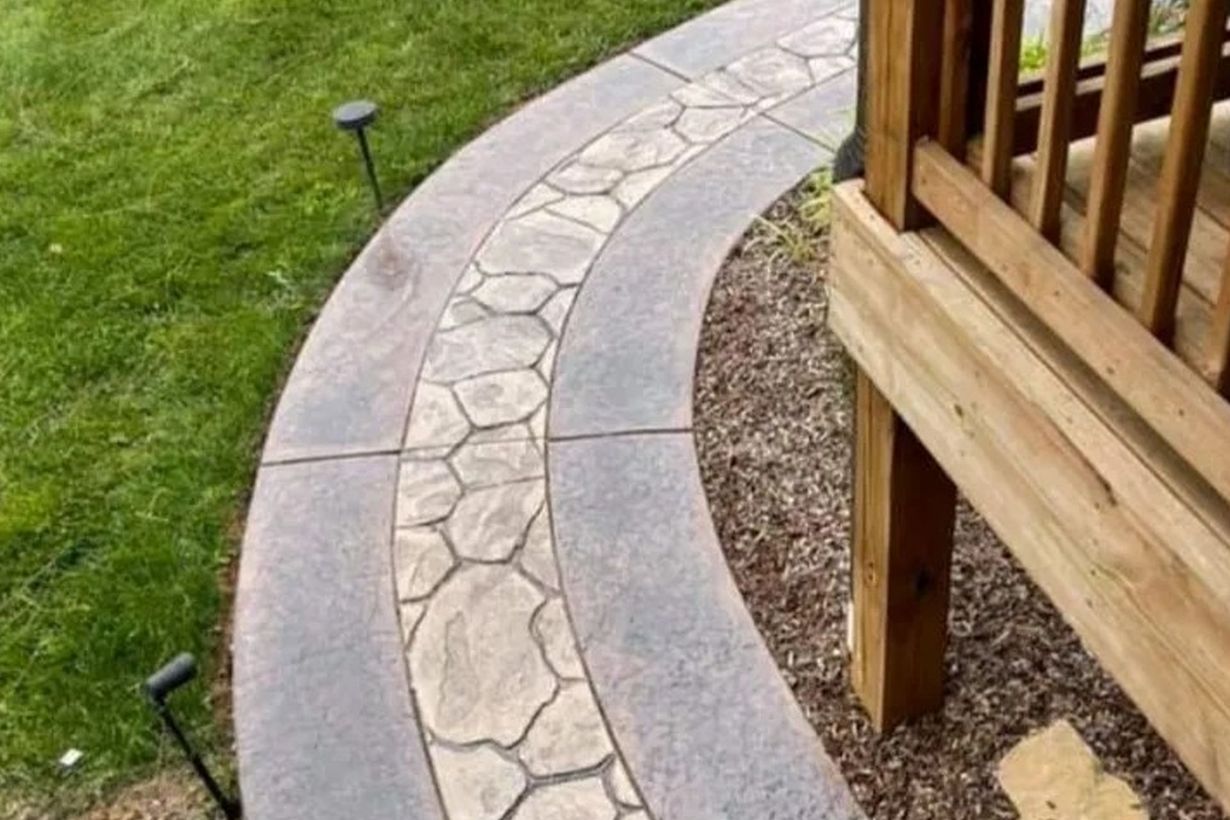 Curving concrete pathway with stone-like center, beside grass and a wooden deck.