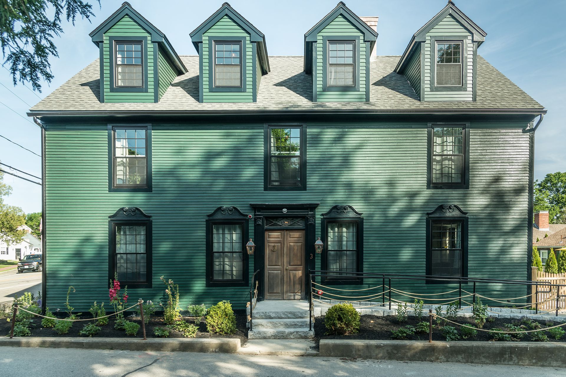 Green historic house with four dormer windows, front porch, and flower beds in bright sunlight
