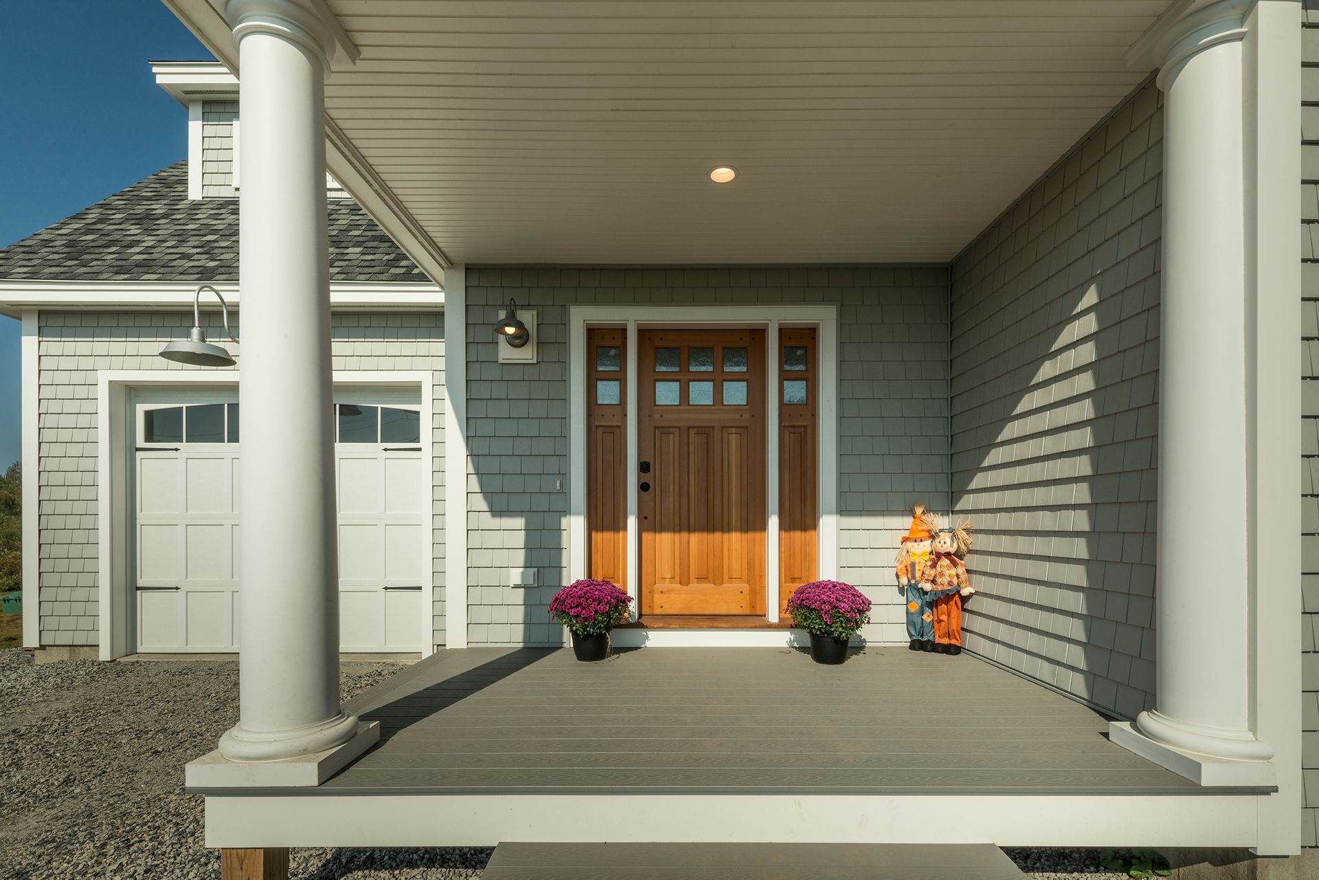 Front porch with white columns, wooden front door, flower pots, and a small autumn scarecrow decoration
