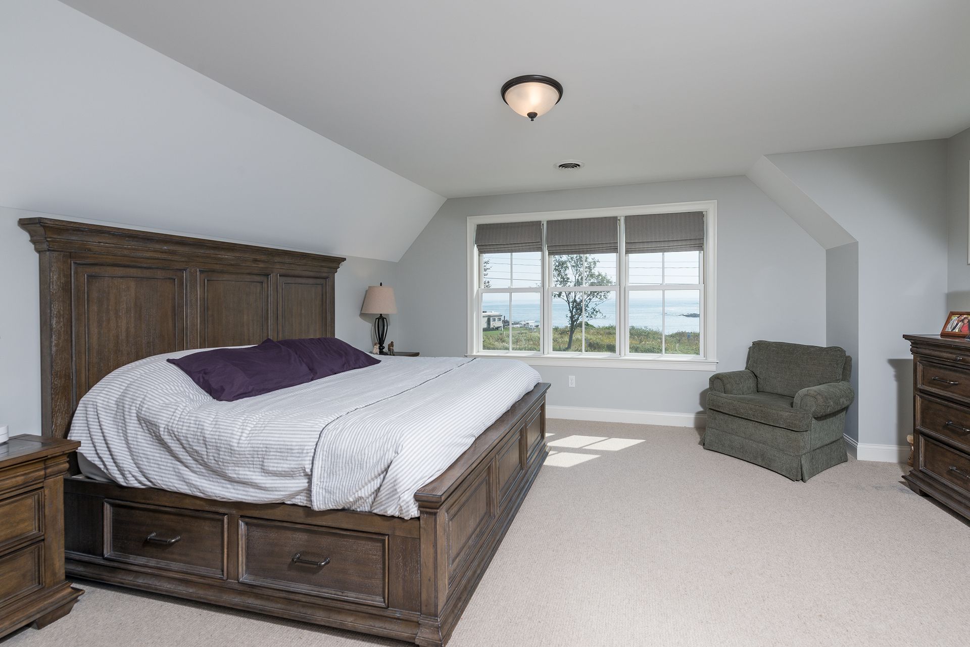 Spacious bedroom with a wooden bed, gray chair, dresser, and large window overlooking trees and mountains
