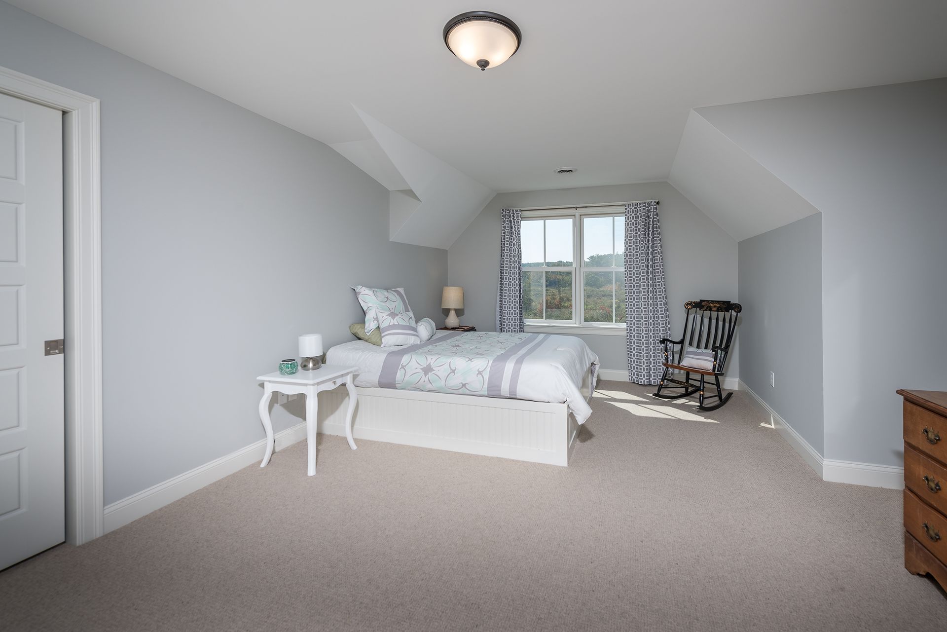 Spacious attic bedroom with a bed, white side table, rocking chair, and window view.