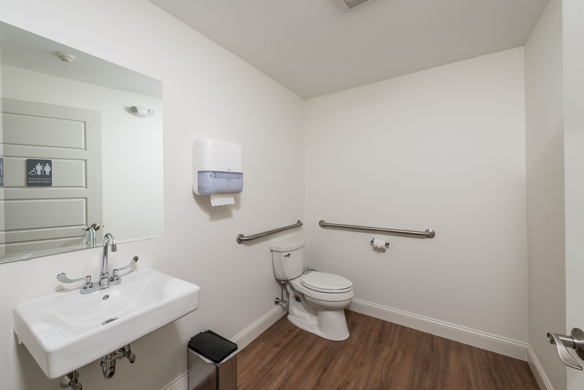 Accessible restroom with sink, toilet, grab bars, and paper towel dispenser, in white walls and wood floor