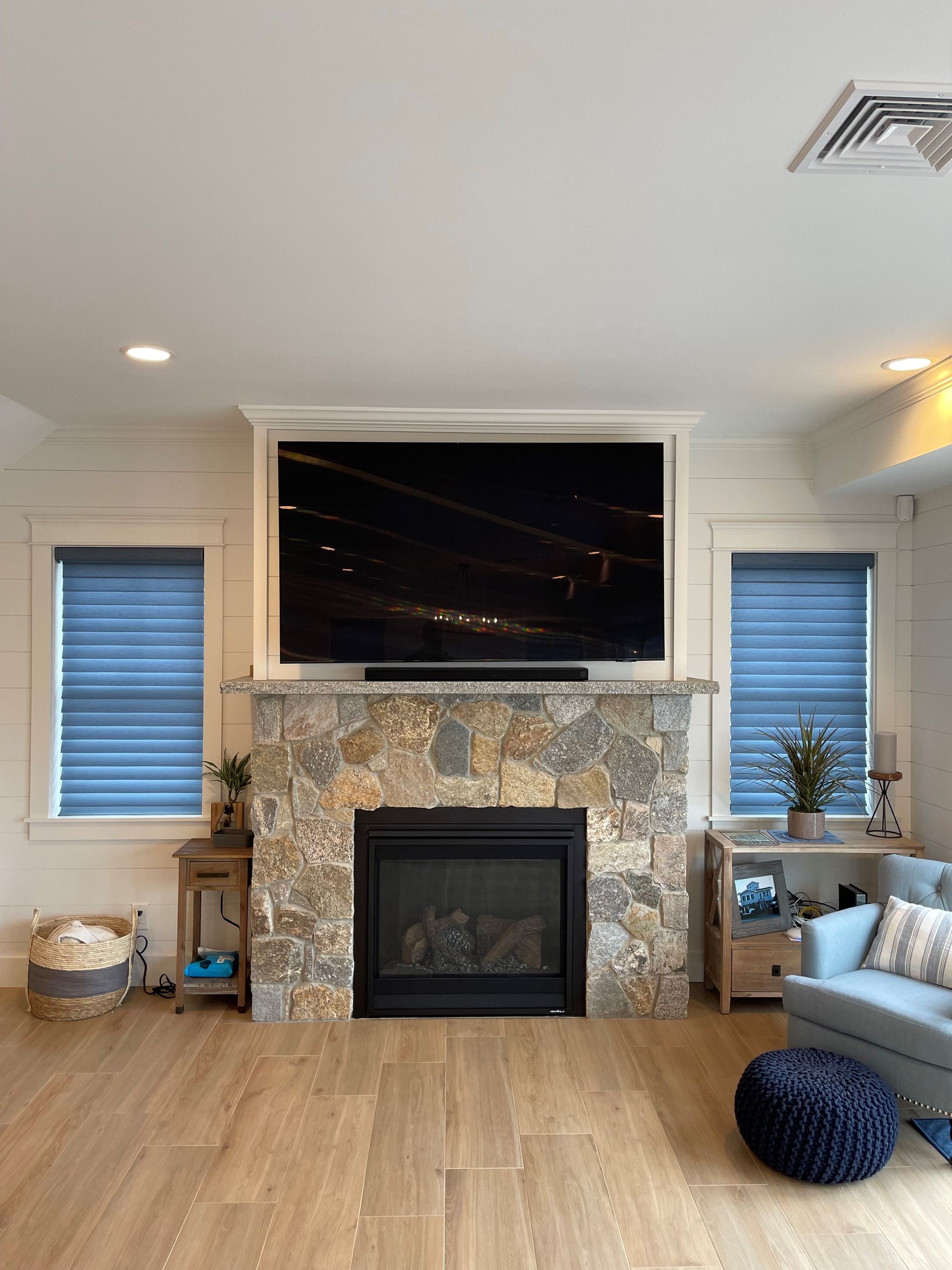 Living room with stone fireplace, wall-mounted TV, blue blinds, and light wood flooring
