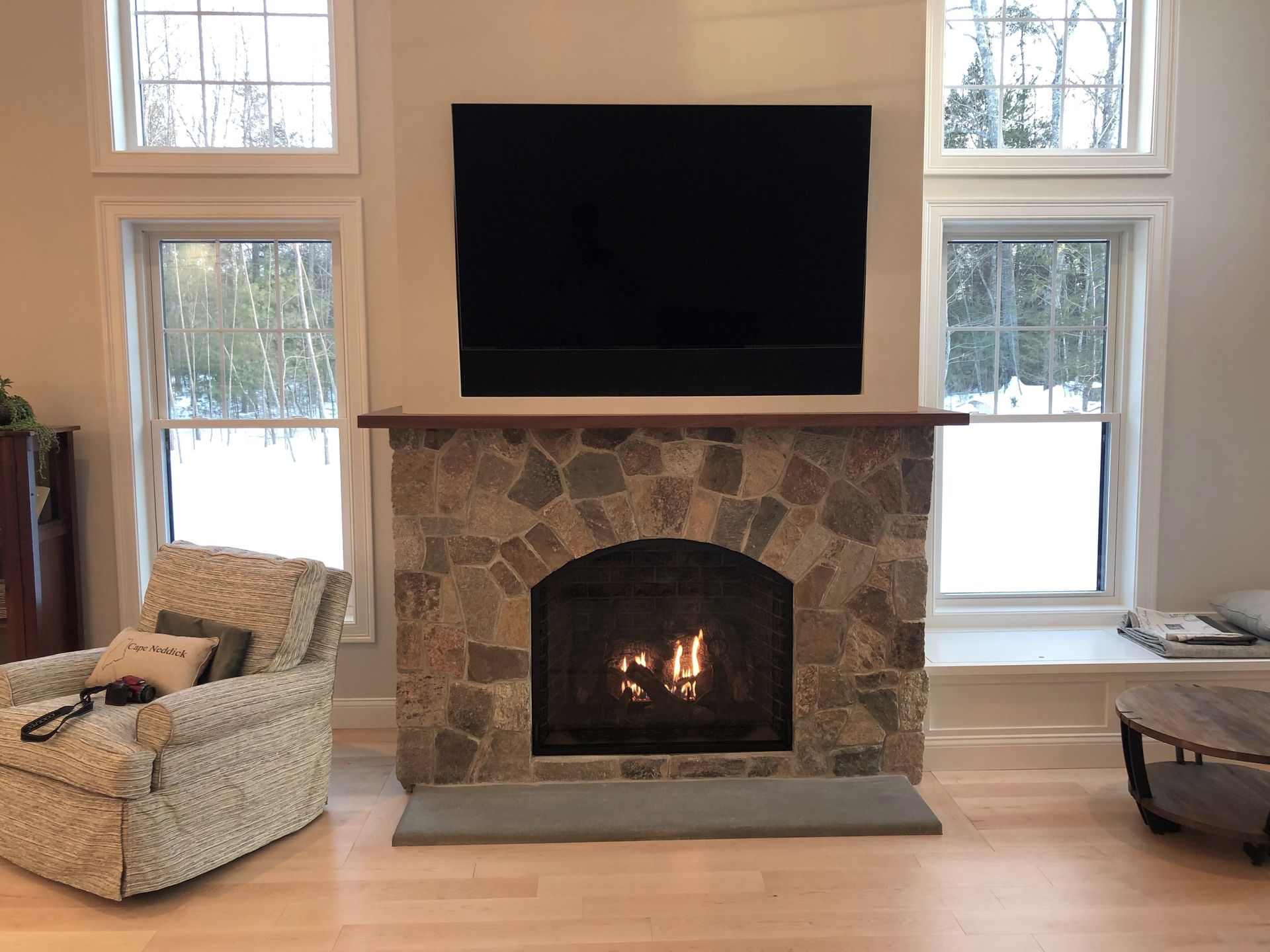 Living room fireplace with a stone surround, wall-mounted TV, and two armchairs by bright windows