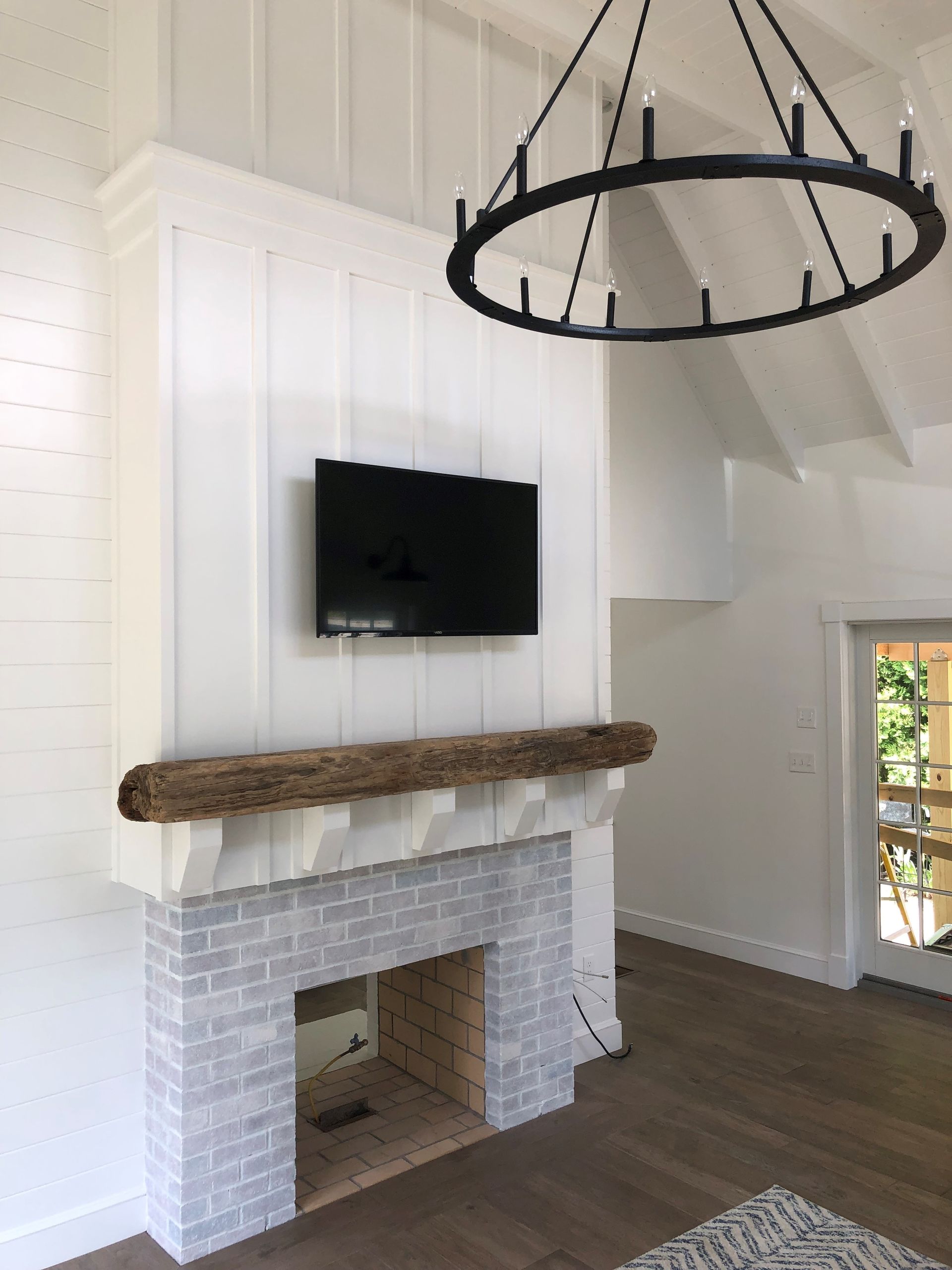 Modern living room with white paneled walls, wall-mounted TV, rustic mantel, and black circular chandelier.