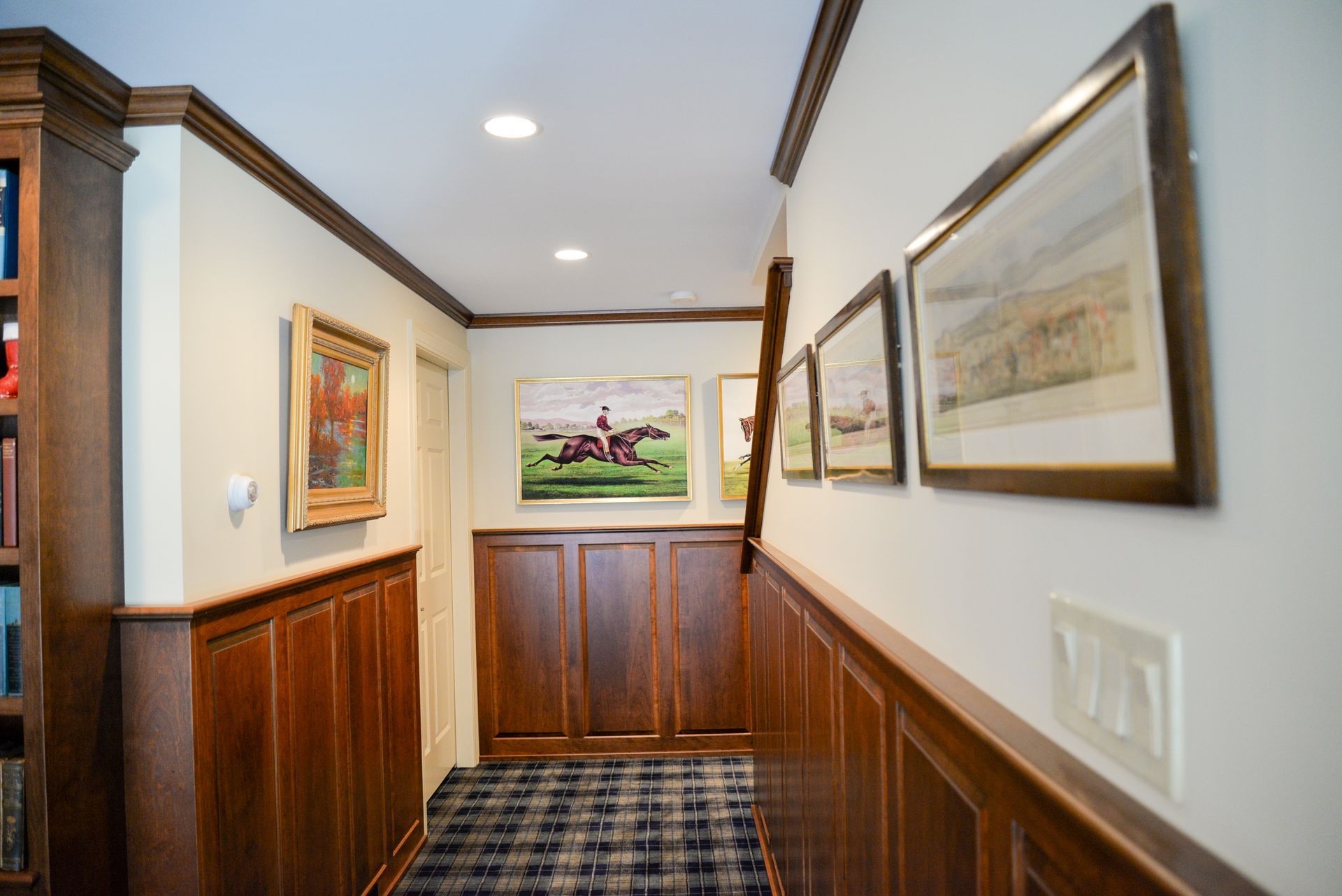 Narrow wood-paneled hallway with framed art, plaid carpet, and recessed ceiling lights