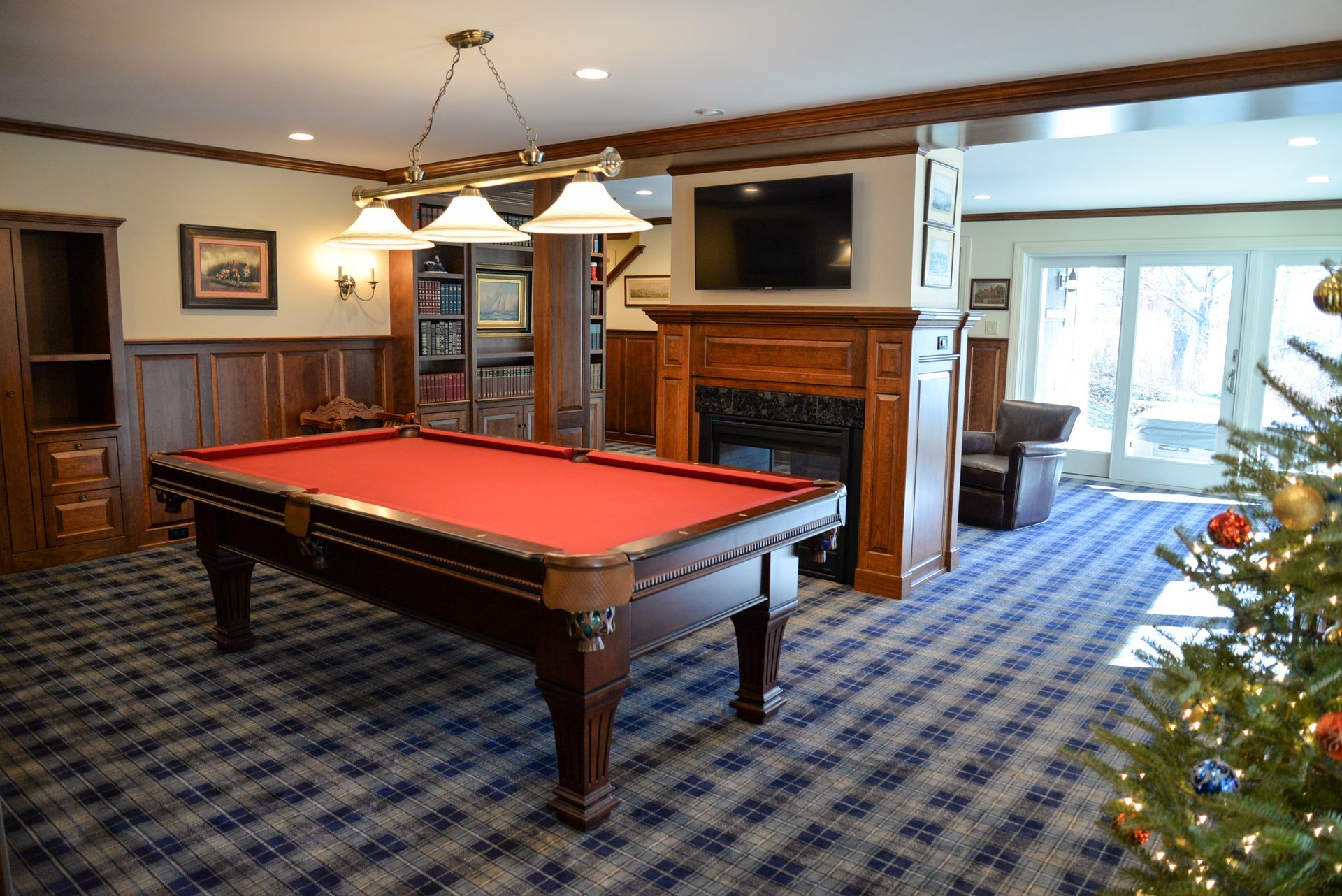 Game room with a red pool table, fireplace, TV, and a lit Christmas tree on blue plaid carpet.