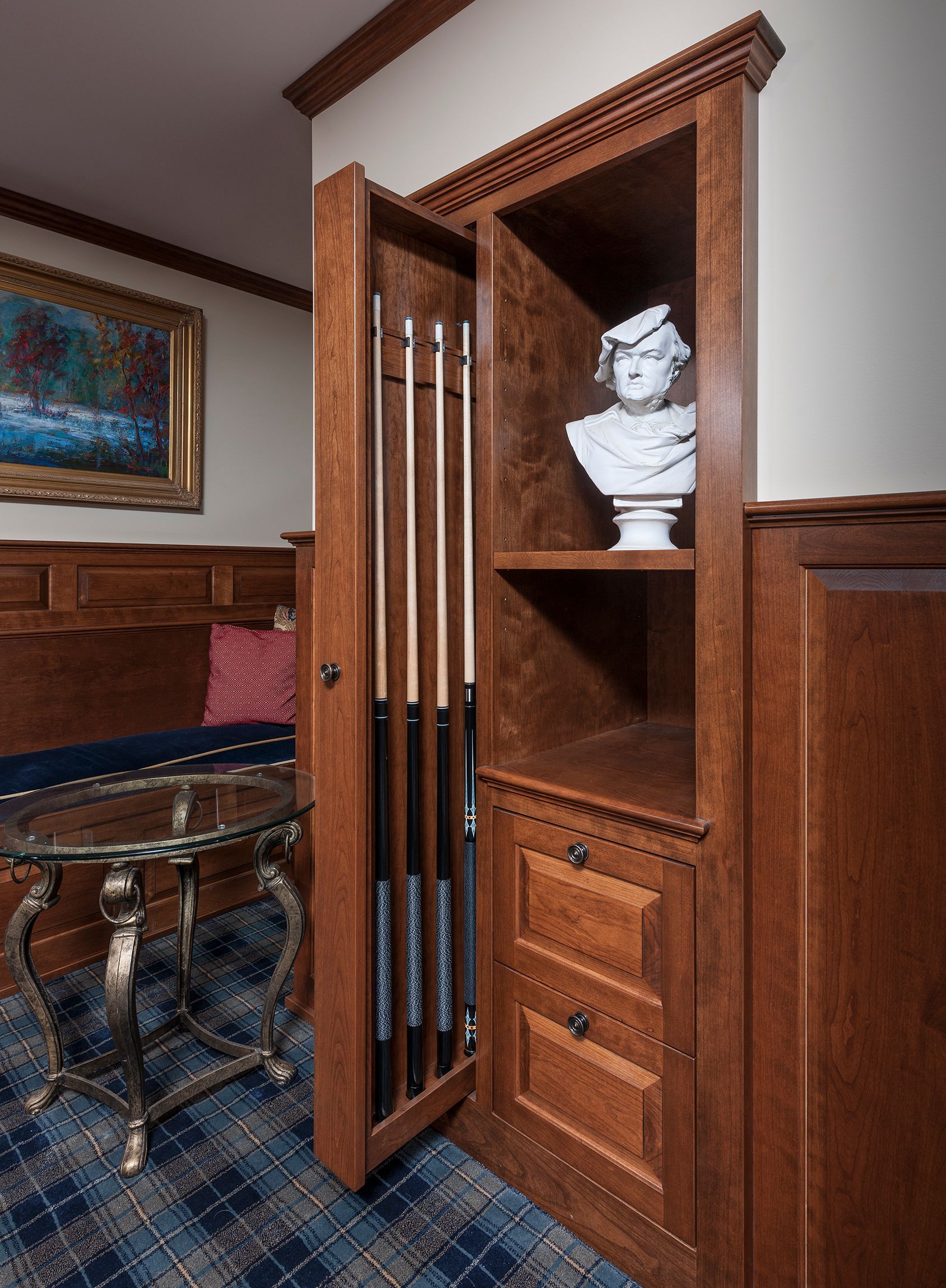 Wooden cabinet with open door revealing shelves, a white bust, drawers, and billiard cues in a room