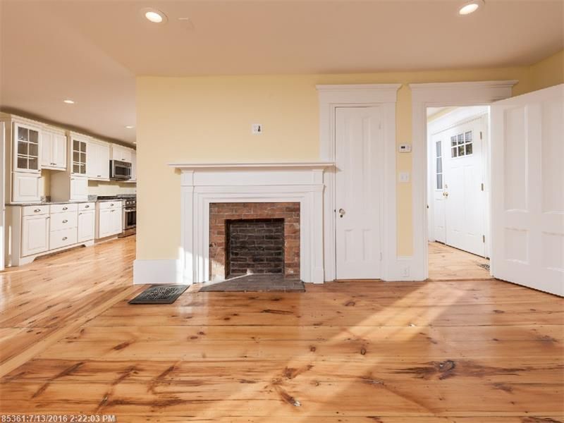 Empty living room with hardwood floors, white fireplace, and open kitchen with white cabinets