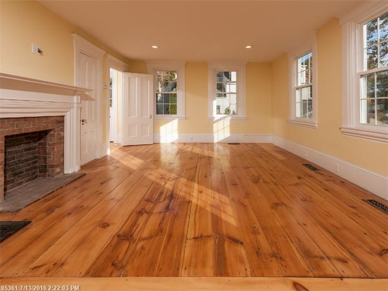 Empty living room with wide plank hardwood floors, white fireplace, and beige walls with tall windows.