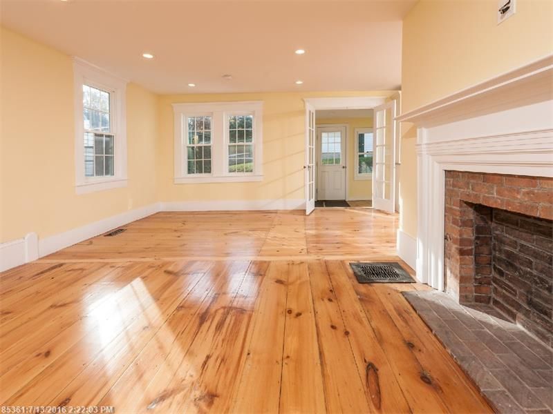 Empty living room with polished hardwood floors, beige walls, windows, and a white fireplace with brick hearth