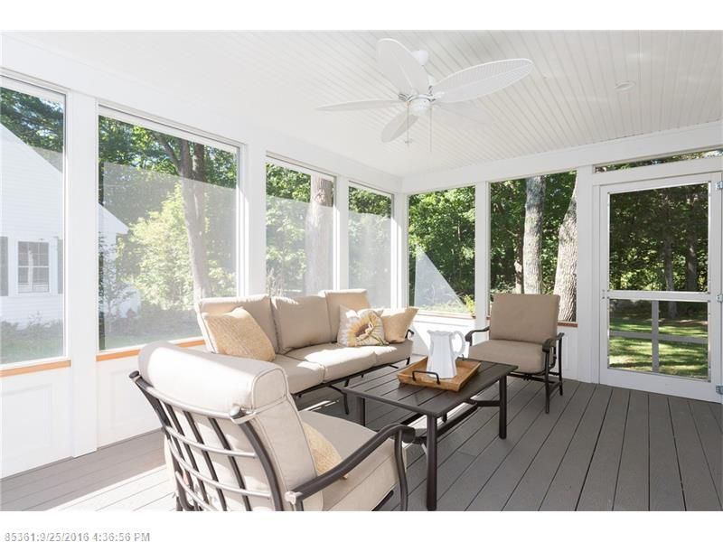Bright screened porch with wicker seating, beige cushions, and a ceiling fan overlooking trees.