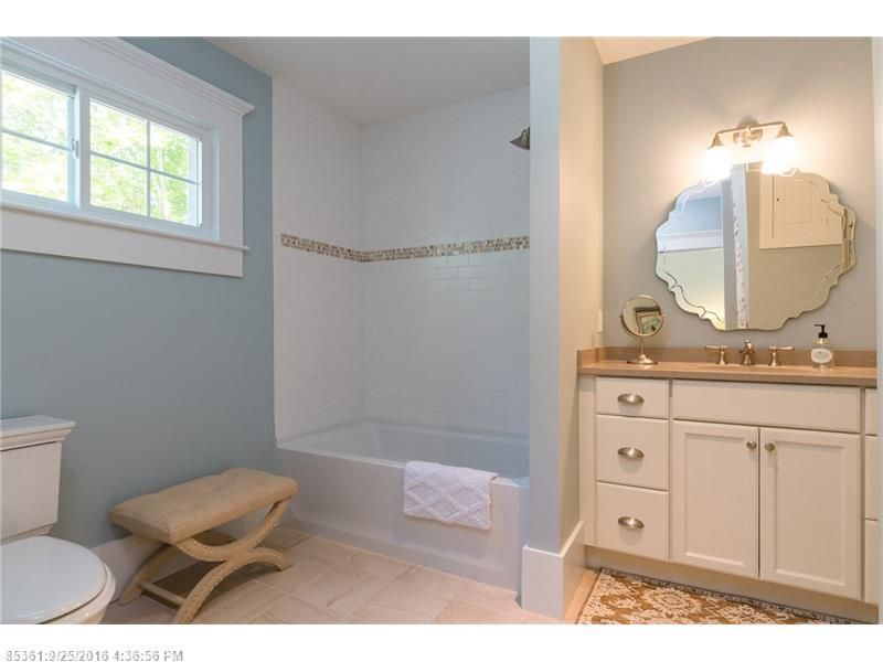 Bright bathroom with tub, window, vanity, and beige ottoman on a tile floor.