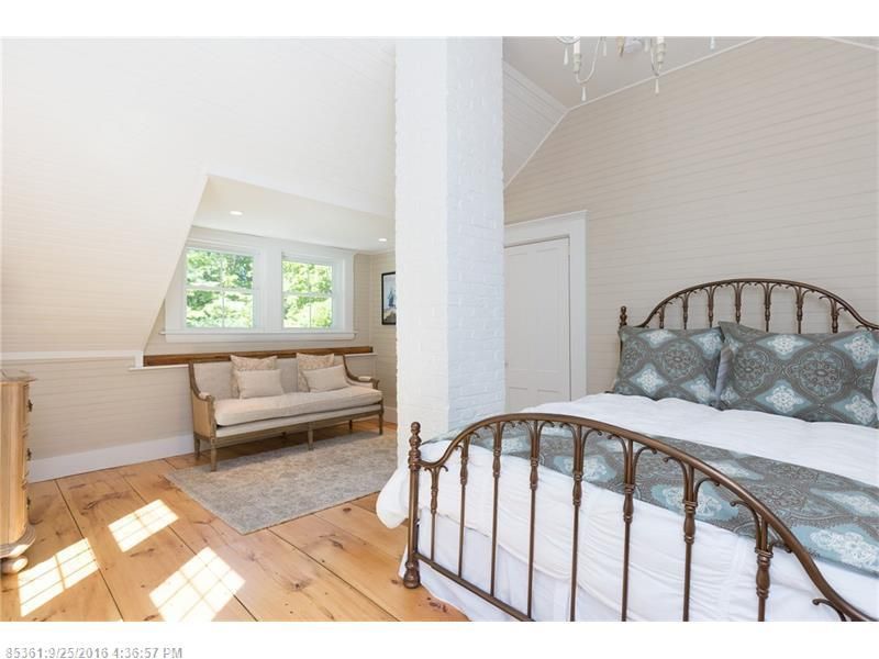 Bright attic bedroom with wrought-iron bed, white walls, wood floor, and a small bench by the window.