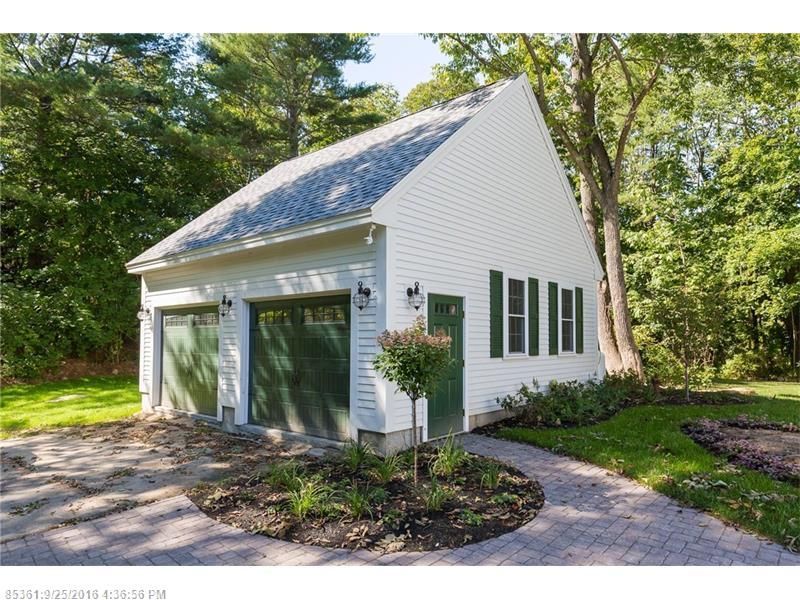 Small white garage with green doors and shutters beside a wooded path, under a tall tree