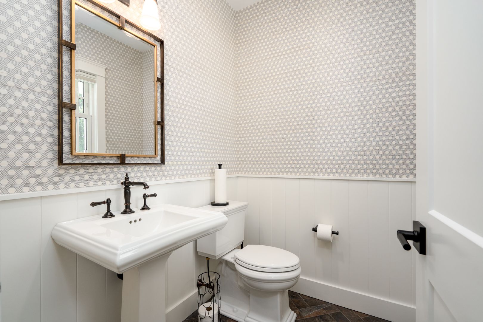 Small bathroom with a white pedestal sink, toilet, mirror, and patterned wallpaper.