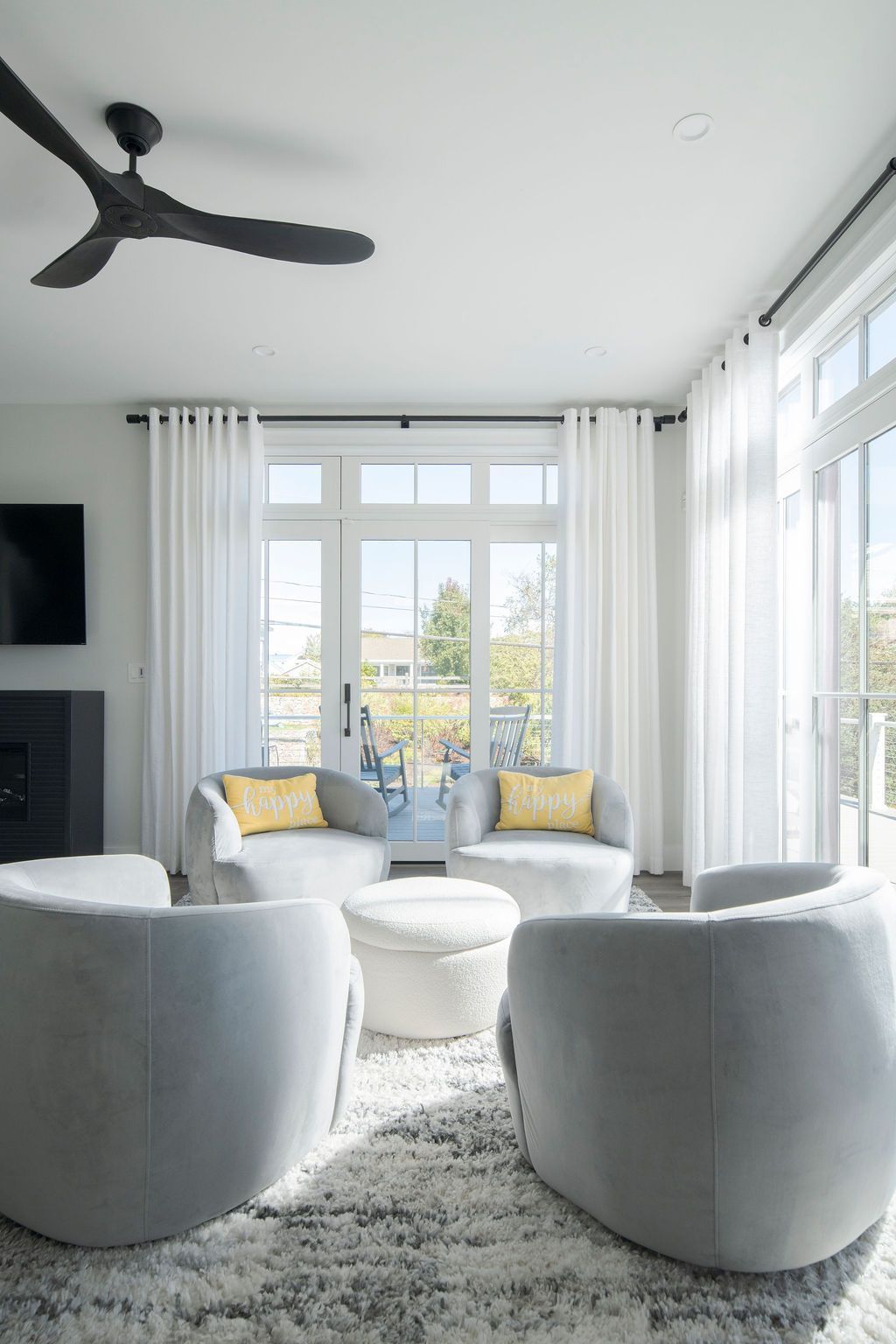 Bright modern living room with white chairs, round ottomans, and large windows overlooking greenery.