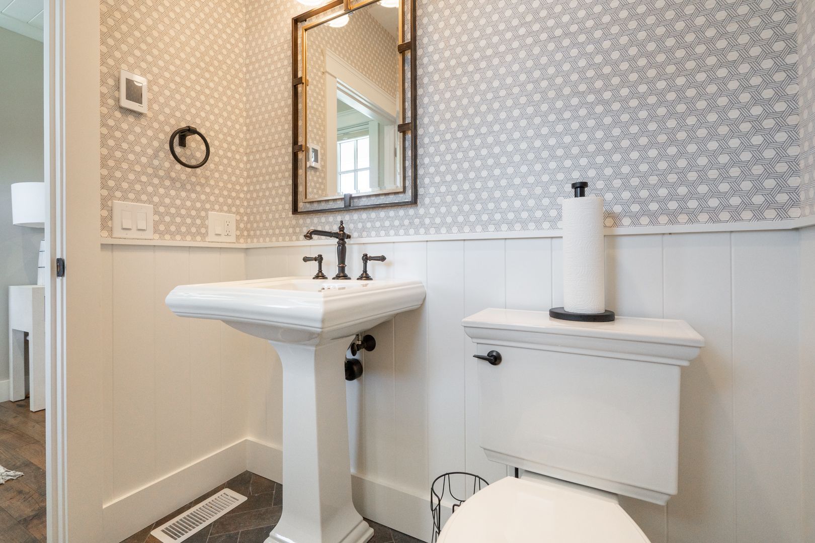 Small bathroom with pedestal sink, toilet, mirror, and patterned wallpaper in neutral tones