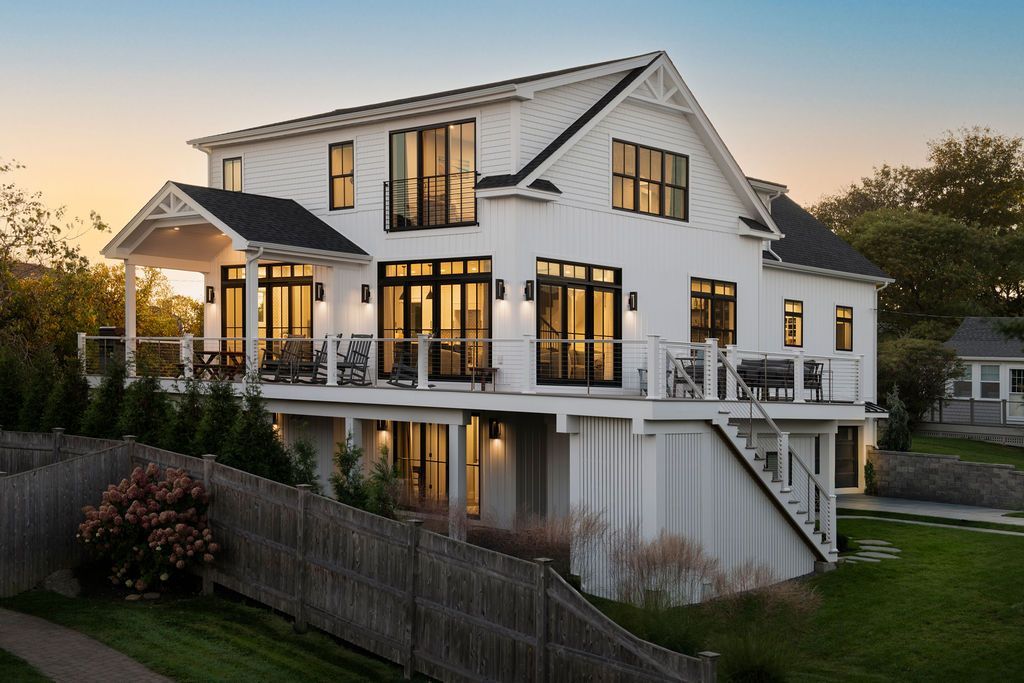 White two-story house with a wraparound porch at sunset, seen above a wooden fence with lit windows.