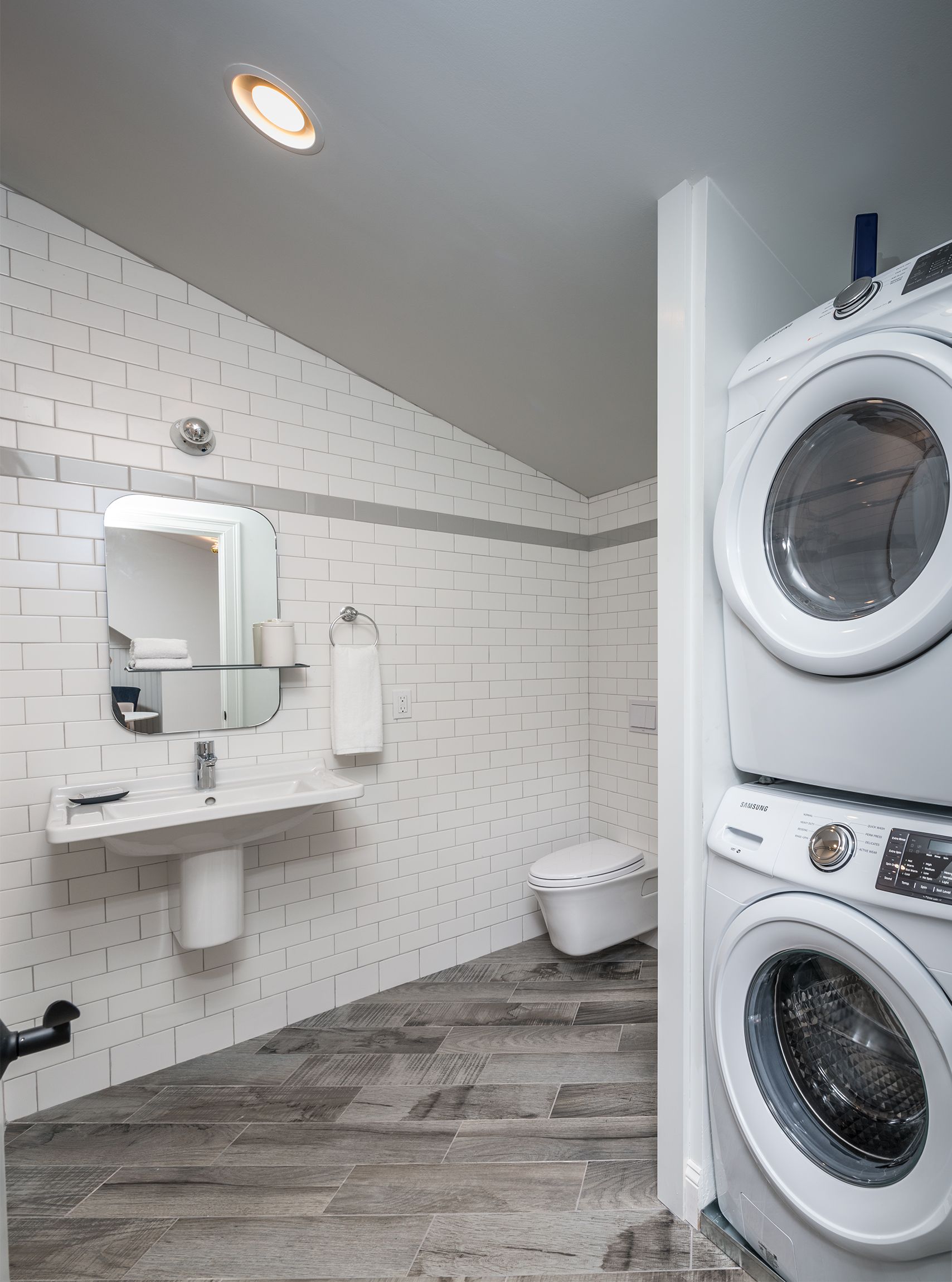 Bright laundry room with stacked washer and dryer, white tiled walls, sink, mirror, and toilet area