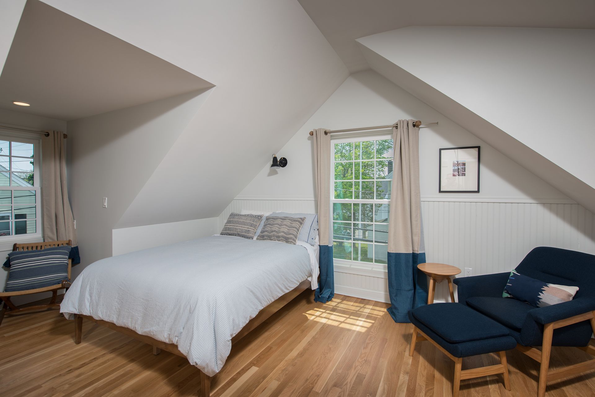 Bright attic bedroom with a white bed, blue chair, wooden floor, and glass door to a balcony.