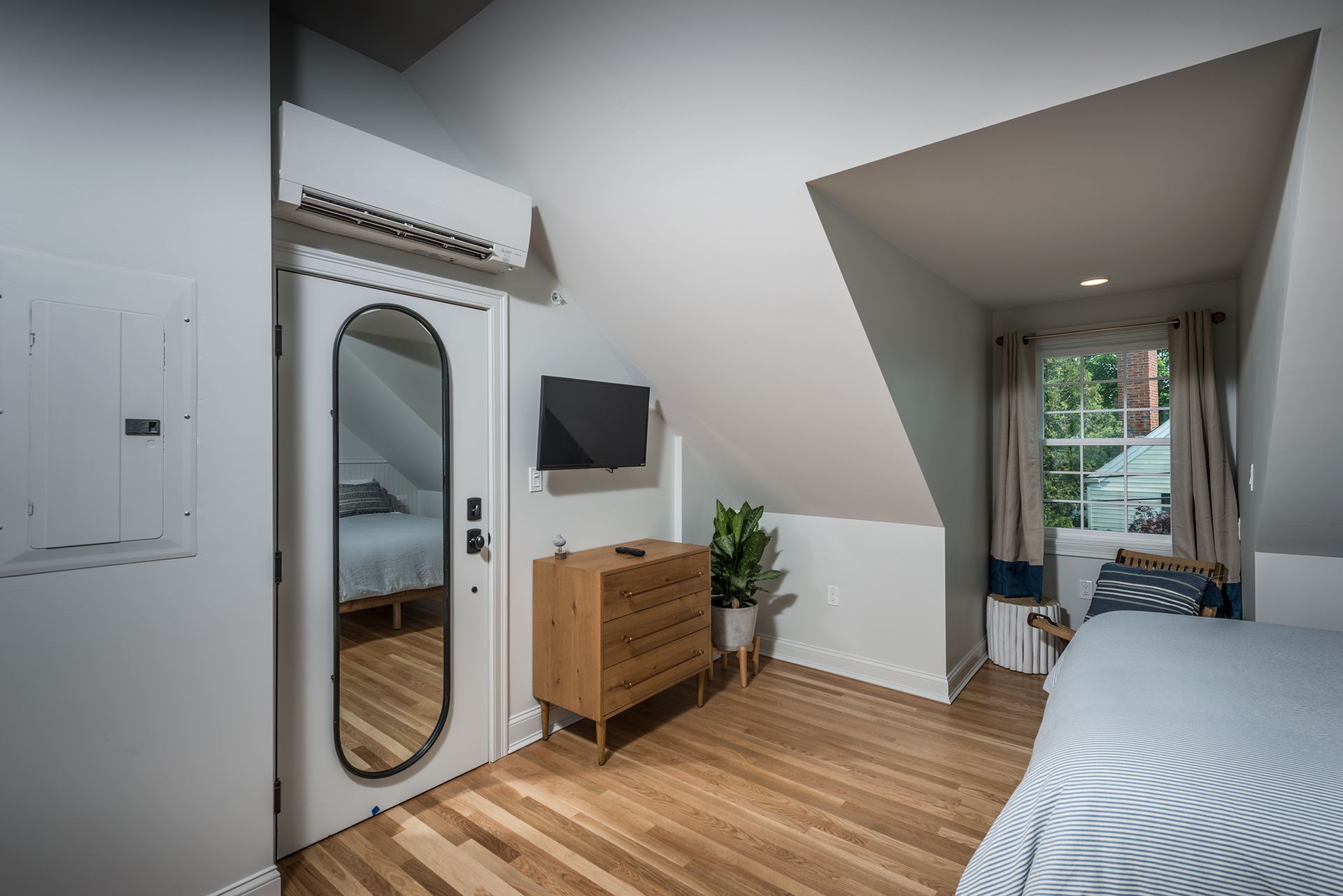 Small attic bedroom with bed, dresser, wall mirror, TV, and sloped ceiling near a windowed doorway.